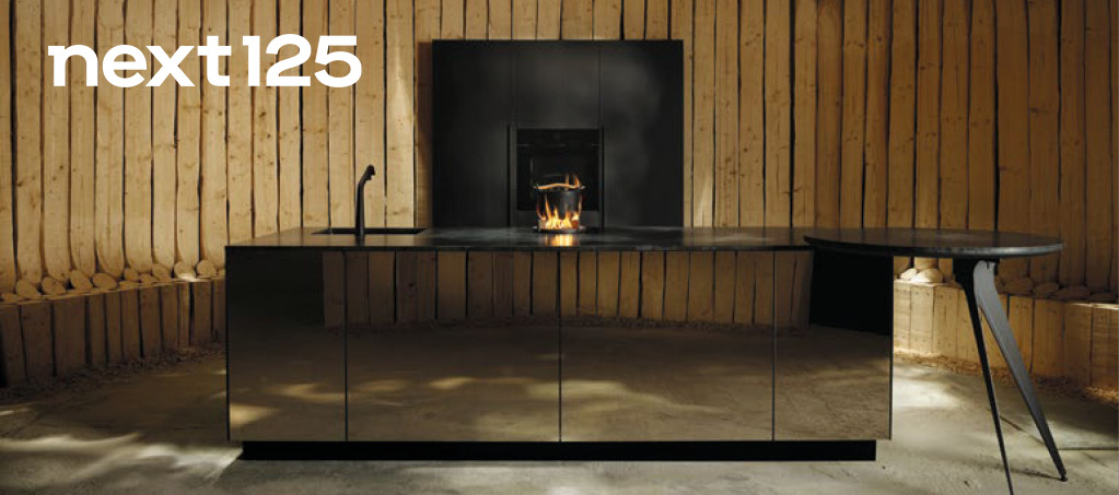 Next125 Küchen bei Himmlisch Wohnen Moderne schwarze Kücheninsel vor Holzverkleidung mit integriertem Kamin