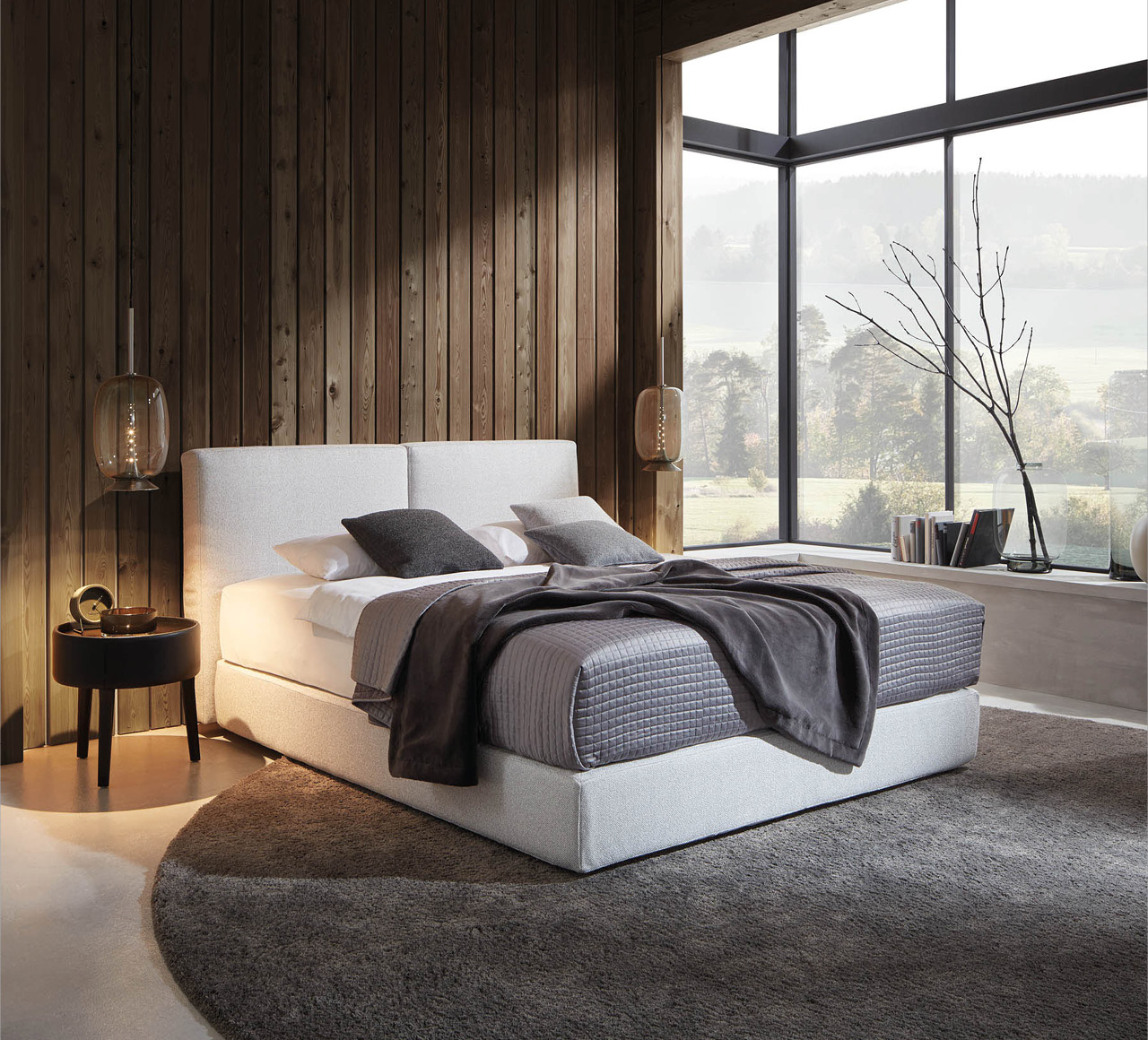 Polsterbett vor Holzvertäfelung und großer Fensterfront mit Ausblick.