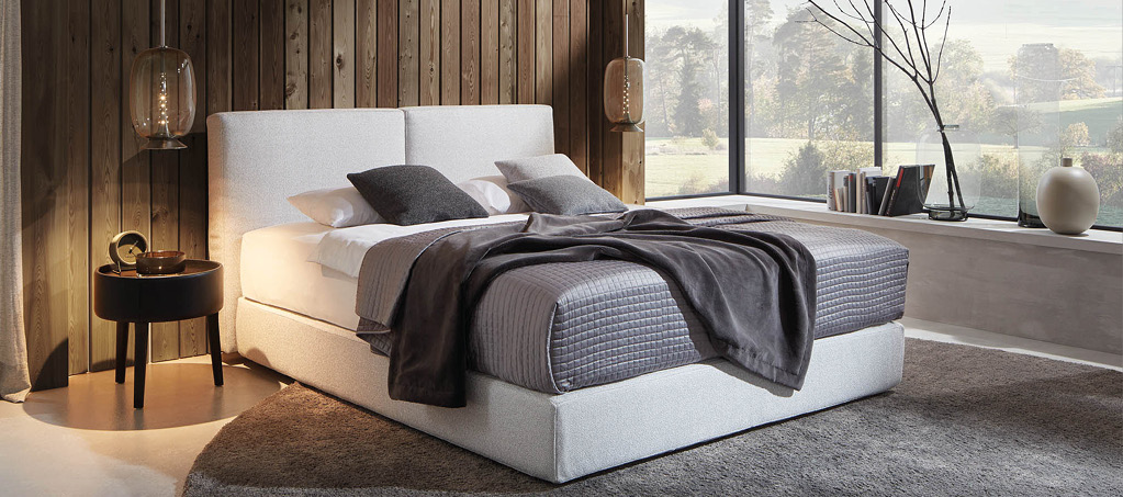 Polsterbett mit Nachttisch vor Holzvertäfelung und Fensterfront.