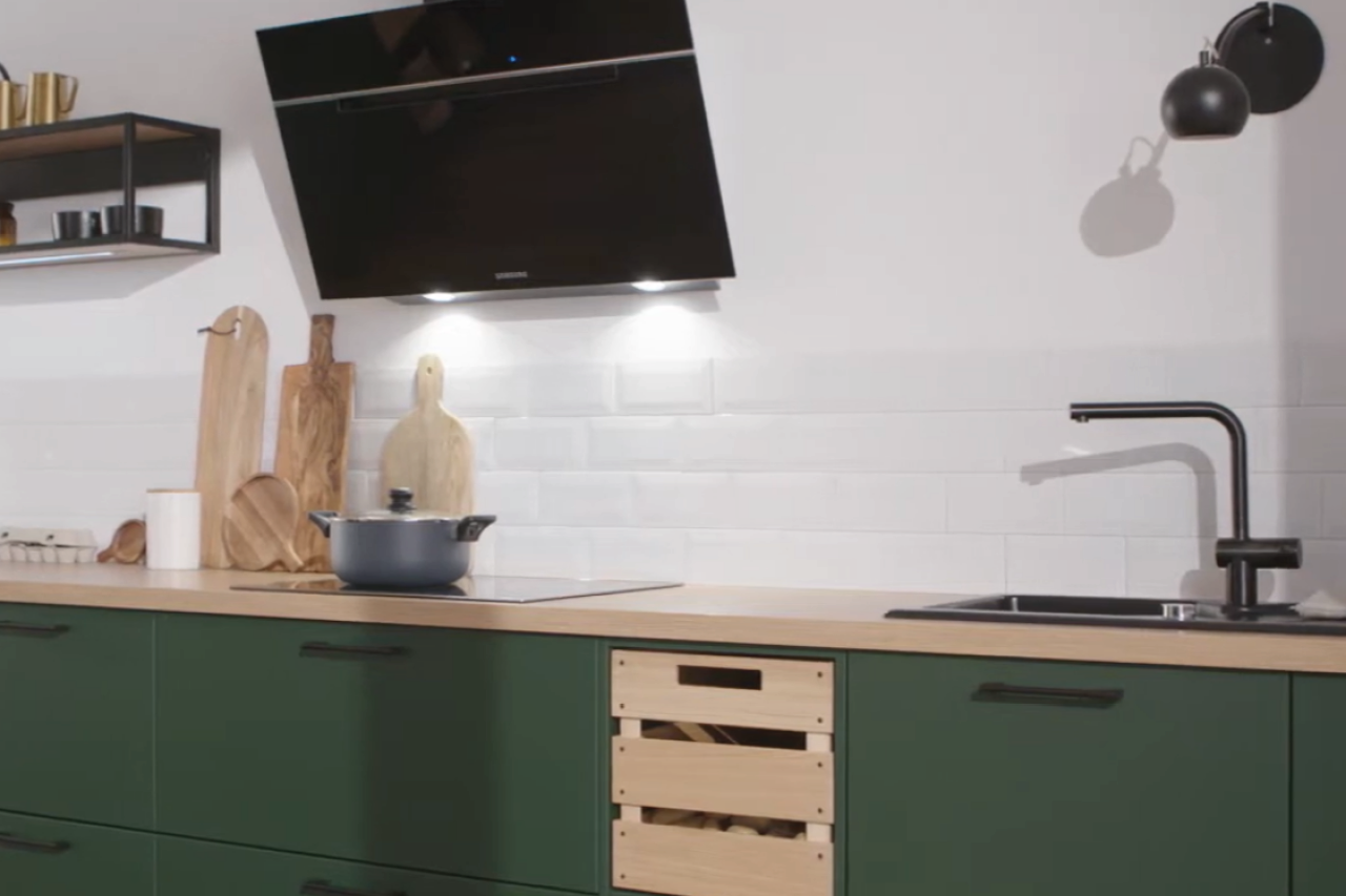 Moderne Einbauküche mit matten grünen Fronten, heller Holzarbeitsplatte und schwarzer Dunstabzugshaube. Zeitgemäßes Küchendesign mit Induktionskochfeld, schwarzer Armatur und weißen Wandfliesen im Hintergrund.