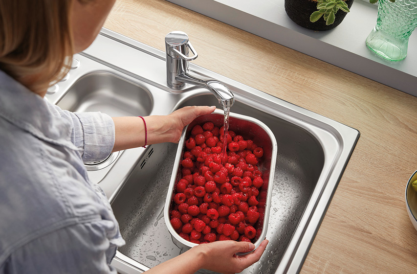 Person wäscht Himbeeren in Schüssel unter Küchenwasserhahn in Edelstahlspüle