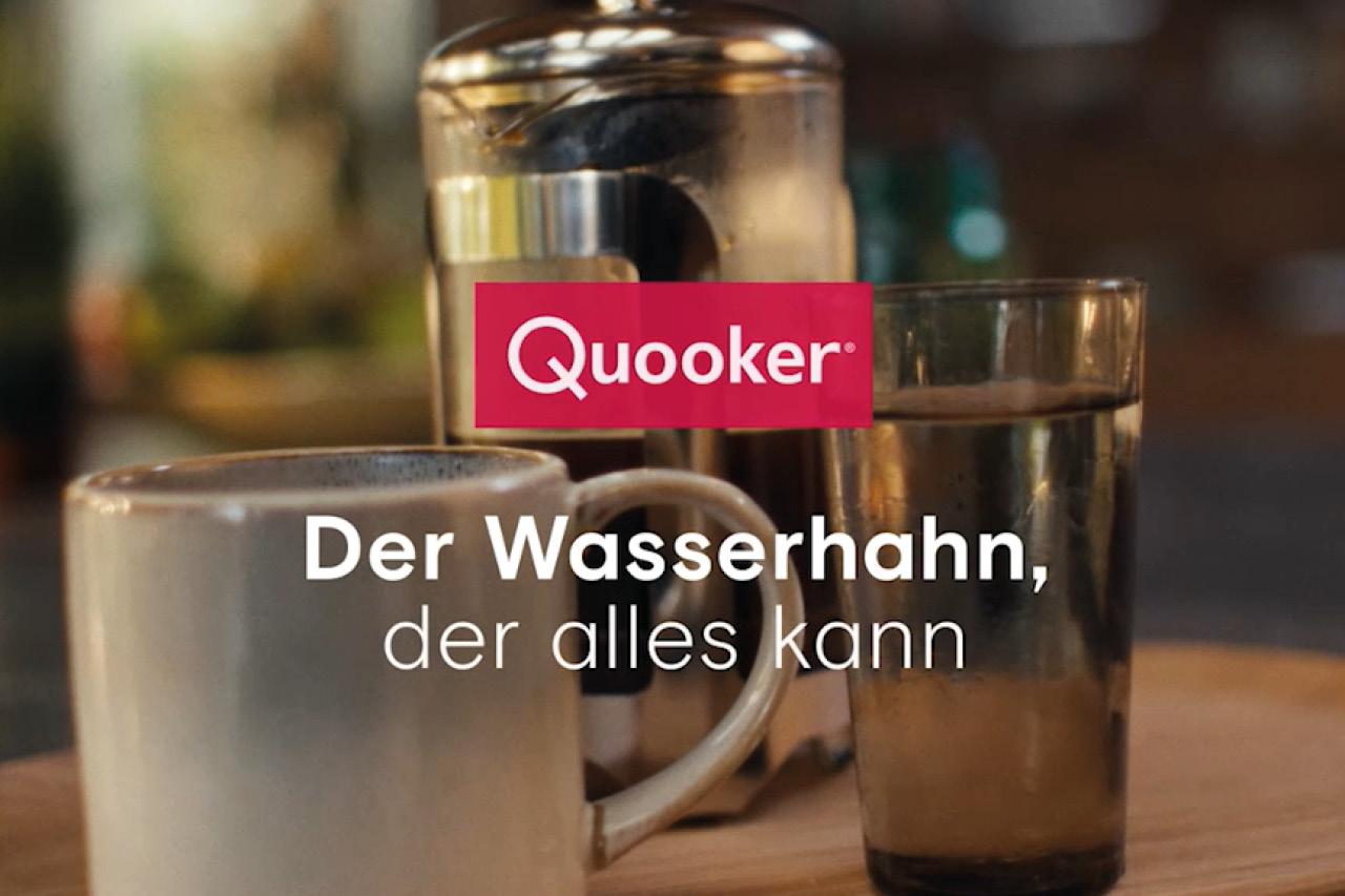 Quooker-Logo und Slogan "Der Wasserhahn, der alles kann" vor unscharfem Hintergrund. 