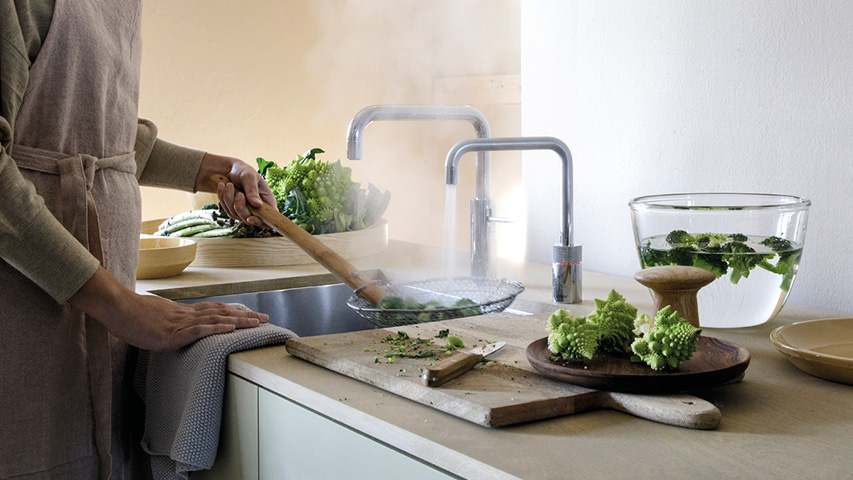 Person blanchiert Gemüse am Quooker Wasserhahn, Dampf über Spüle.