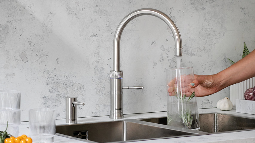 Quooker Wasserhahn füllt Glasgefäß am Spülbecken, Kräuter im Wasser.