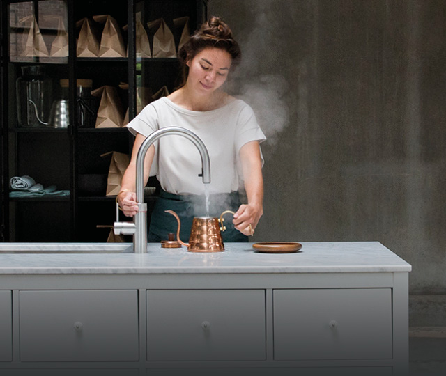Person nutzt Küchenarmatur mit Dampf über Kanne, Link zu Quooker