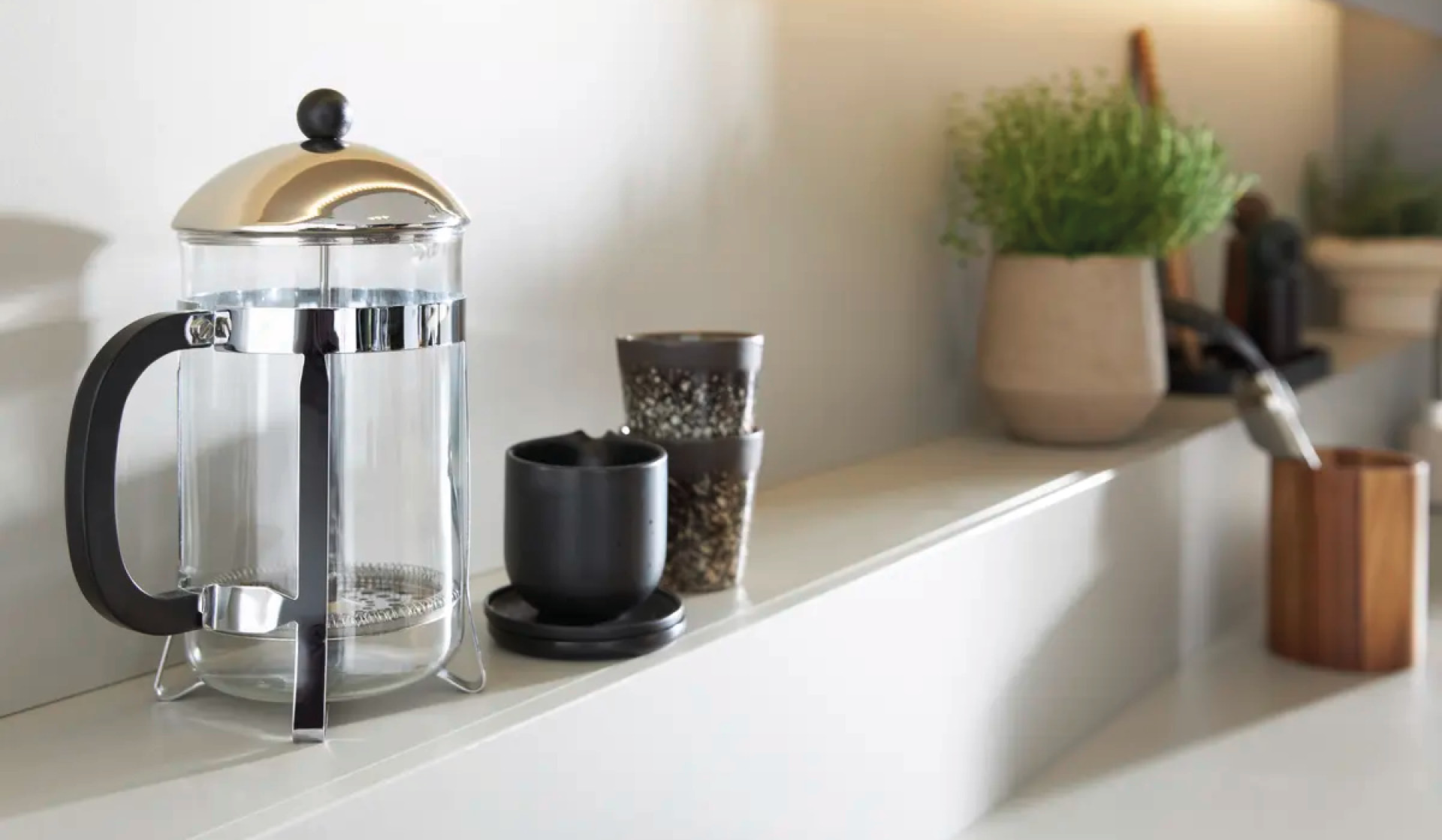 Kaffeezubehör auf Küchenablage mit French Press, Tassen und Pflanze.
