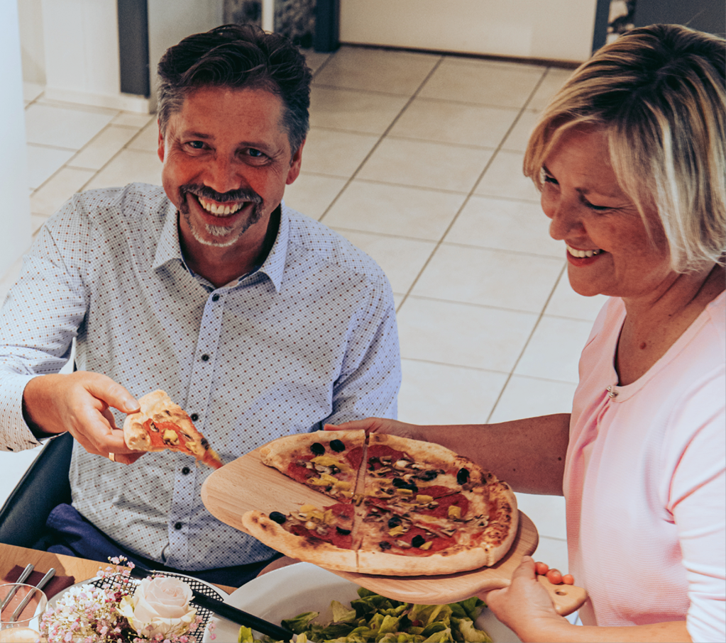 Mann und Frau sitzen am Tisch, halten Pizzastücke und ein Holzbrett mit Pizza, Salat und Geschirr auf dem Tisch