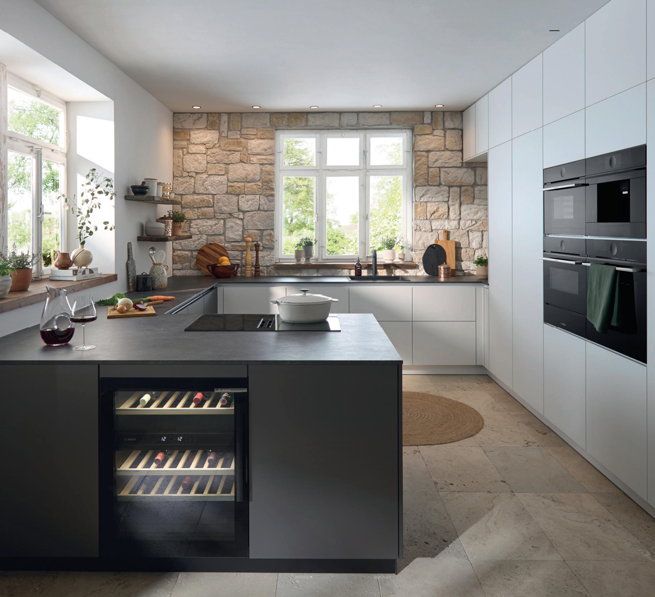 Küche mit weißer Front, Kochinsel mit Kochfeld und Weinkühler, Steinwand hinten und Fenster mittig