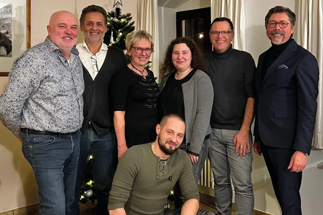 Mehrere Personen stehen und sitzen zusammen vor einem Weihnachtsbaum in einem Raum mit Vorhängen