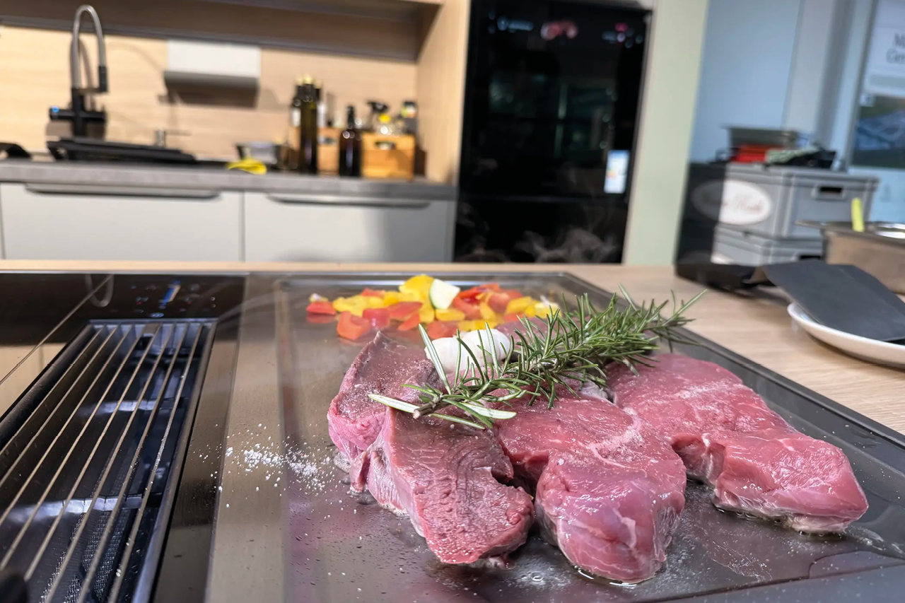 Stücke Fleisch mit Kräutern liegen auf Kochfeld, geschnittenes Gemüse dahinter, Küchenzeile im Hintergrund