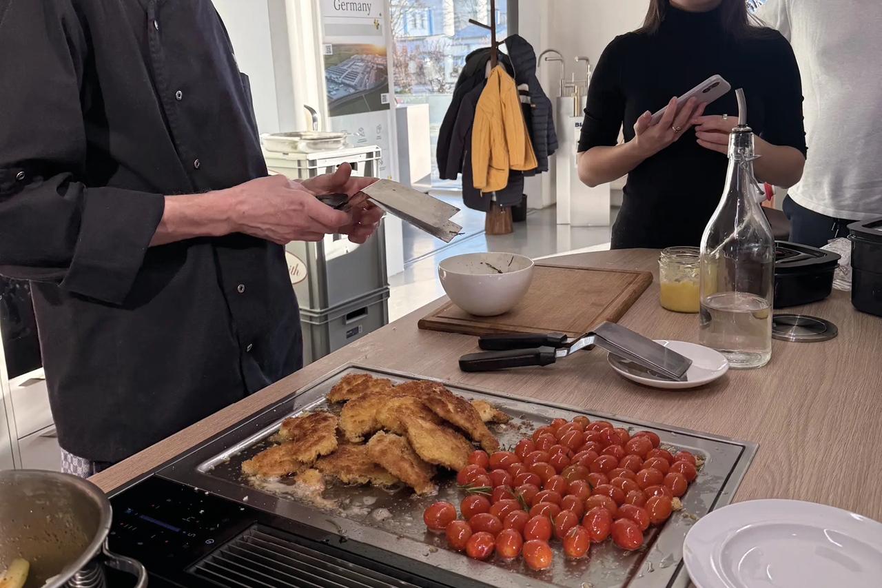 Panierte Stücke und Tomaten liegen auf Kochfeld, Person mit Messer und Schneidebrett daneben, Küche im Hintergrund