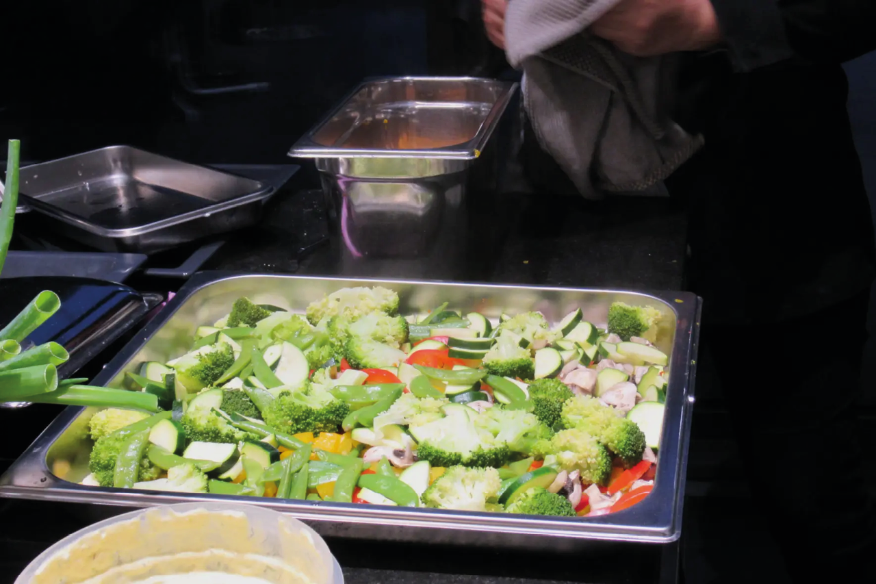 Edelstahlbehälter mit Brokkoli, Paprika und Zucchini auf Herd, Küchenutensilien und Behälter im Hintergrund