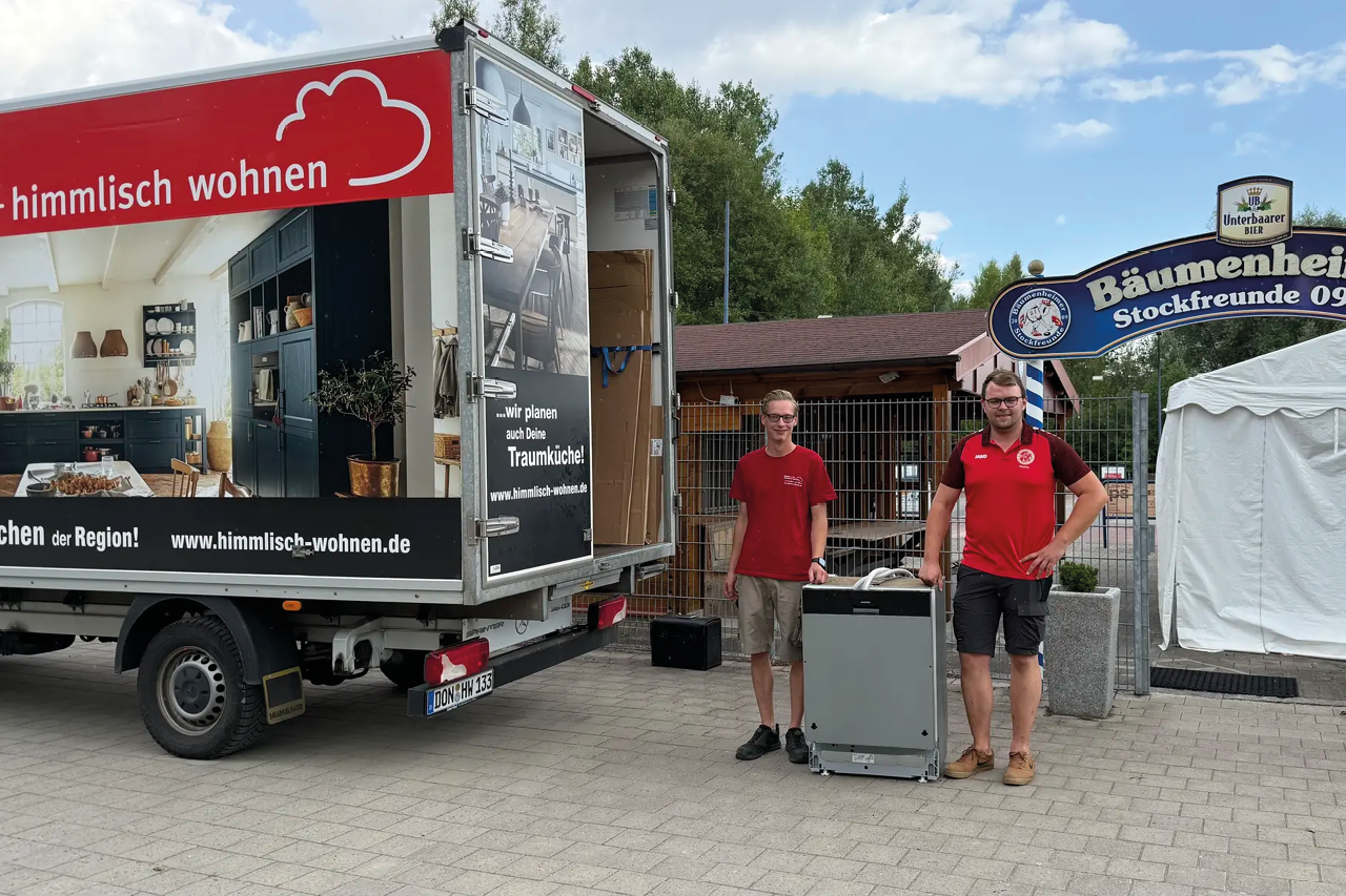 Lkw mit Aufdruck himmlisch wohnen steht vor Eingangstor, zwei Personen daneben mit Gerät auf Boden