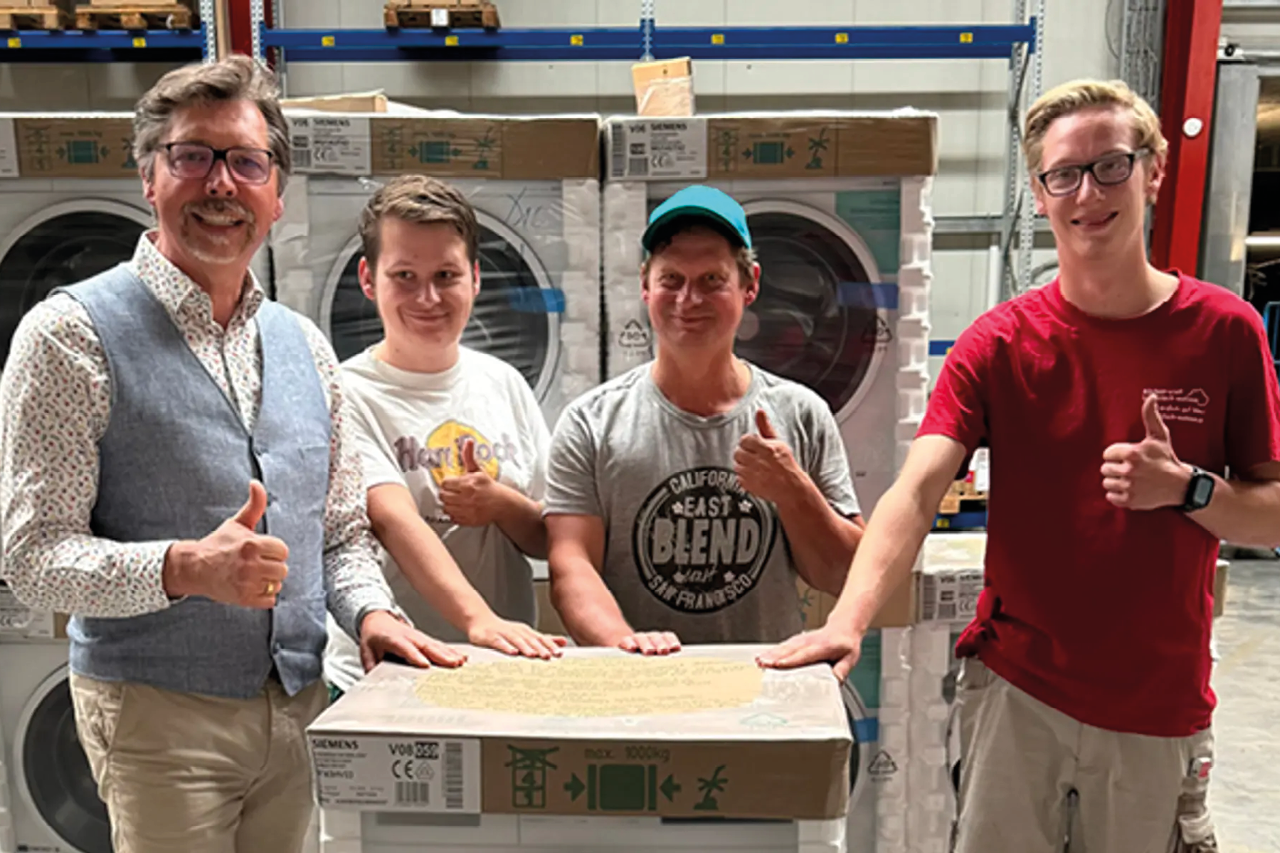 Vier Personen stehen vor verpackten Geräten in Lager, zeigen Daumen nach oben und halten Karton vorne