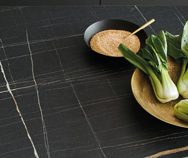 Dunkle Küchenarbeitsplatte aus Naturstein oder Keramik mit feiner Maserung. Auf der Arbeitsfläche liegen frisches Gemüse und eine Schale mit Getreide. Symbolbild für hochwertige Küchenarbeitsplatten und moderne Küchenausstattung.