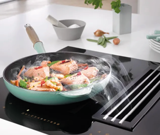 Pfanne mit Lachs und Gemüse auf einem modernen Induktionskochfeld mit integriertem Kochfeldabzug. Dampf wird direkt abgesaugt. Innovative Küchentechnik für offene Wohnküchen.