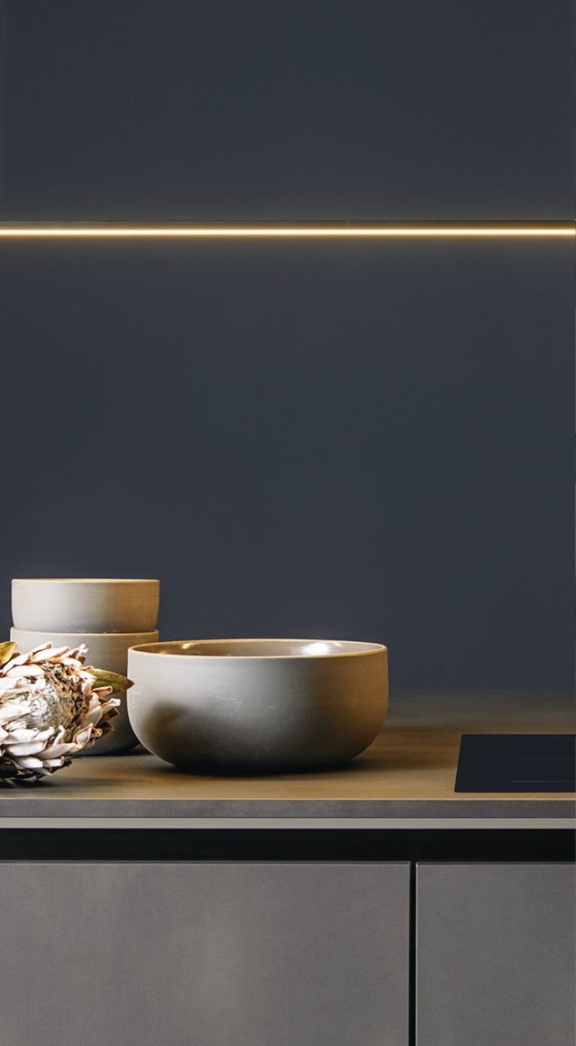 Minimalistische Küchenzeile mit dunklen Fronten, integrierter LED Lichtleiste und Keramikschalen auf der Arbeitsplatte. Moderne Designküche mit klaren Linien und hochwertiger Küchenplanung.
