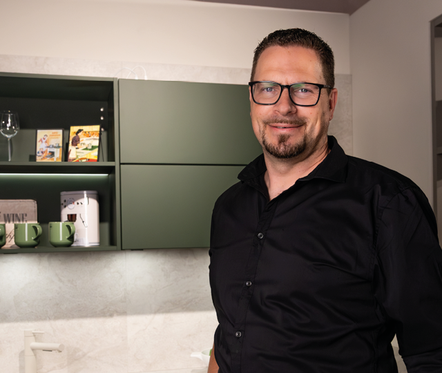 Mann mit schwarzem Hemd steht vor Küche mit grünen Oberschränken, offenen Fächern und Tassen im Regal