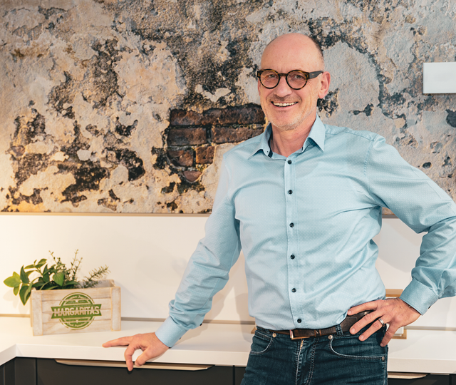 Mann mit Brille steht vor Wand mit großem Bild, Küchenarbeitsplatte und Holzkiste mit Pflanze