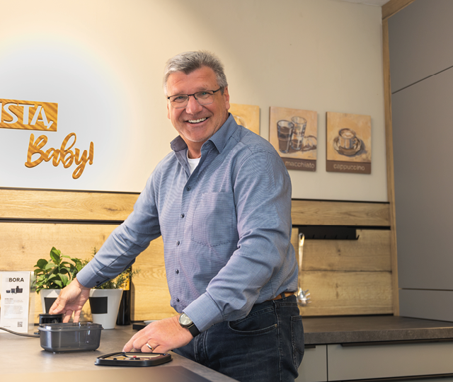 Mann steht an Arbeitsplatte mit Kochfeld und Topf, Holzrückwand und Bildern mit Kaffee-Motiven im Hintergrund