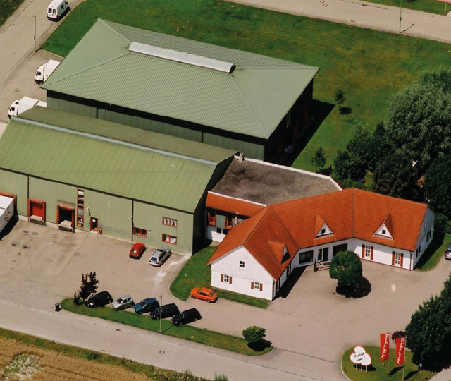 Luftbild mit mehreren Gebäuden, Hallen mit grünem Dach und Wohnhaus mit rotem Dach, Fahrzeuge davor