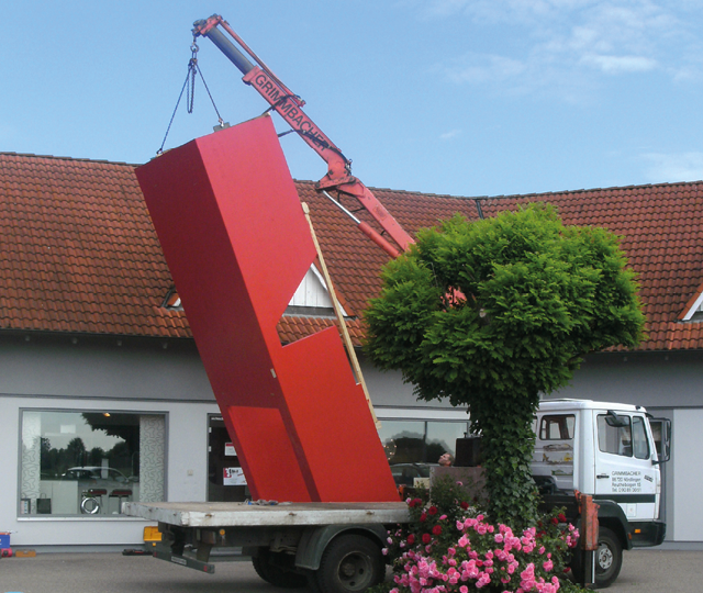 Kran hebt großes rotes Bauteil über Lkw vor Gebäude mit rotem Dach und Baum im Vordergrund