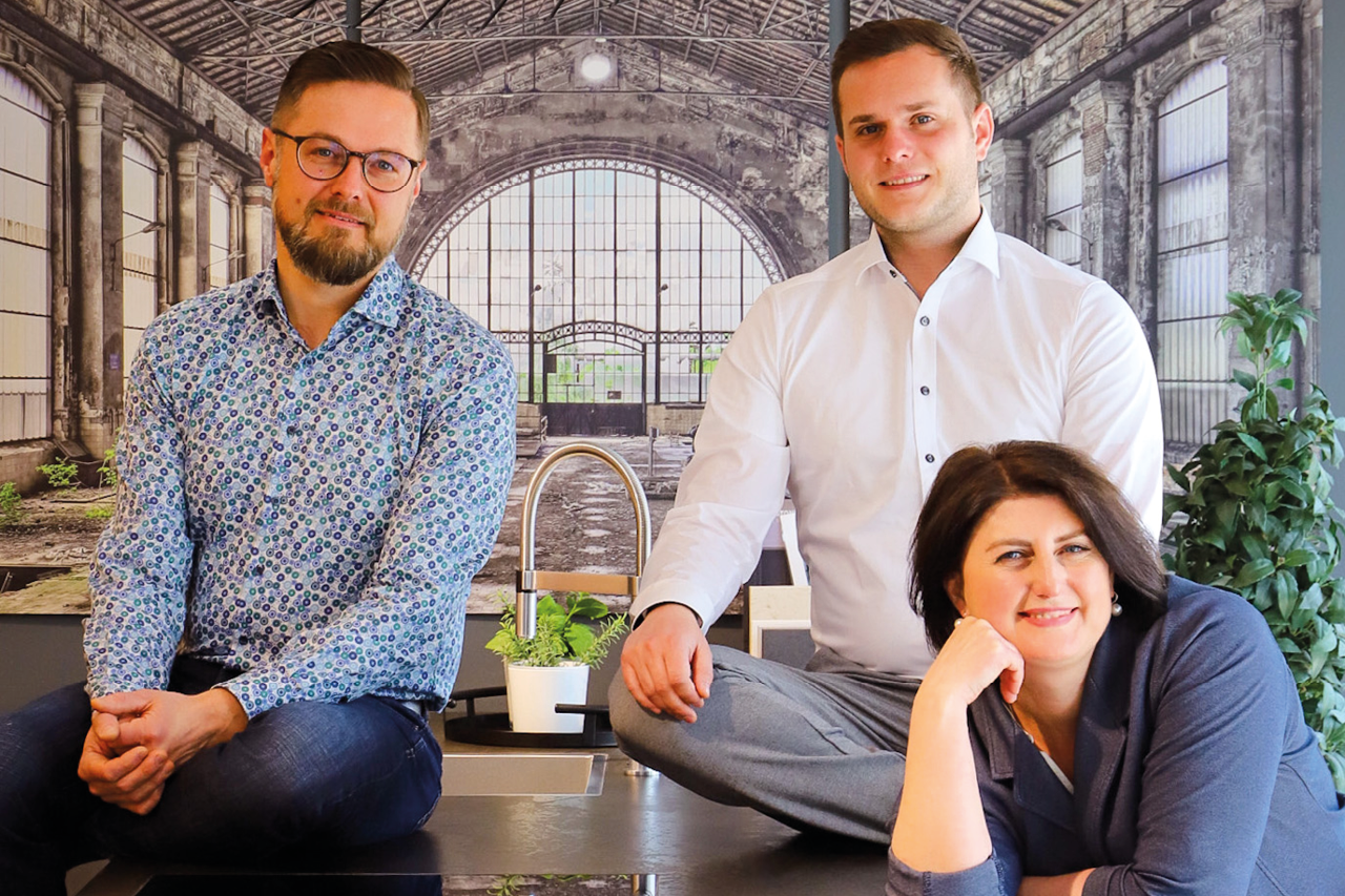 Drei Personen sitzen und stehen an Kücheninsel, Spüle und Pflanze davor, große Wand mit Fensterstruktur im Hintergrund