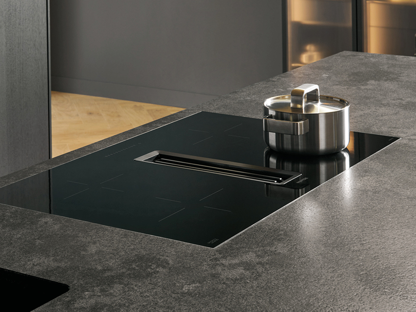 Modernes Induktionskochfeld mit integriertem Muldenlüfter in dunkler Kücheninsel. Hochwertige Arbeitsplatte in Steinoptik und minimalistische next125 Design Küche mit innovativer Kochfeldtechnik.