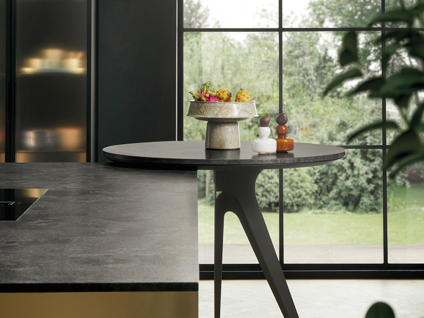 Moderne Kücheninsel mit dunkler Arbeitsplatte und integriertem Ansetztisch vor großer Fensterfront. Design Küche von next125 mit fließendem Übergang zwischen Kochen und Wohnen.