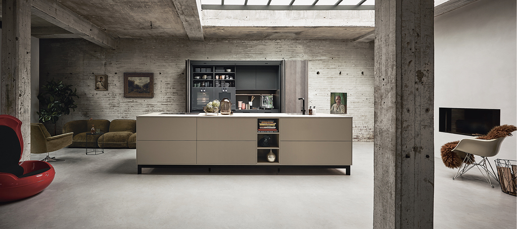 Große Wohnküche im Loft mit zentraler Kochinsel, im Hintergrund offene Regale, links Sofa und Sessel, rechts Kamin und Stuhl