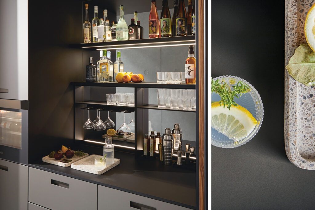 Offener Schrank mit beleuchteten Regalen, gefüllt mit Flaschen, Gläsern und Barzubehör, rechts Glas mit Zitrone auf Arbeitsfläche