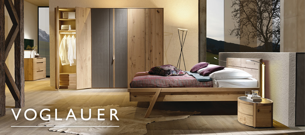 Schlafzimmer mit Massivholzbett, Schrank und „Voglauer“-Schriftzug im Header.