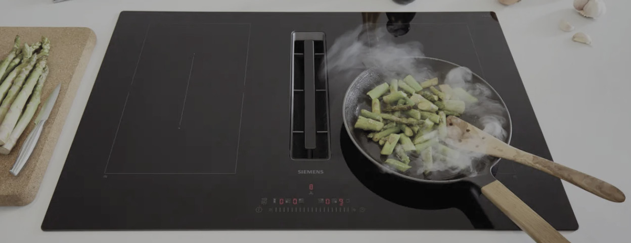 Kochfeld mit integrierter Lüftung und Pfanne mit Gemüse und Dampf, Spargel auf Schneidebrett links