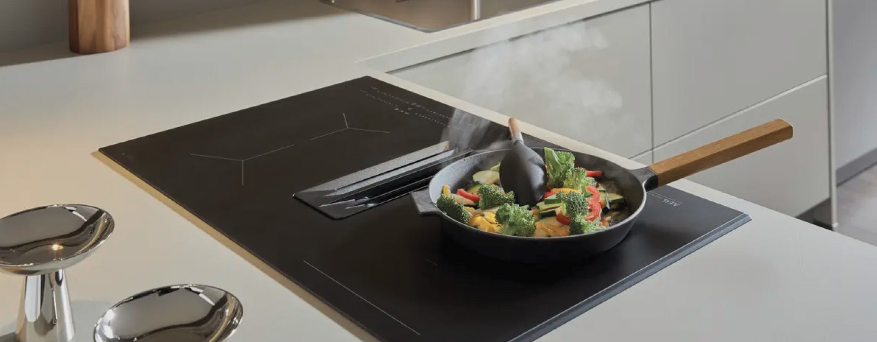 Kochfeld mit integrierter Lüftung, Pfanne mit Gemüse und Dampf, zwei Hocker links am Tresen