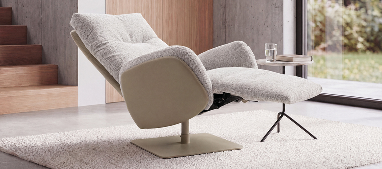 Relaxsessel mit Hocker auf Teppich Heller Relaxsessel mit Hocker, kleinem Tisch und Fensterfront vor Betonwand